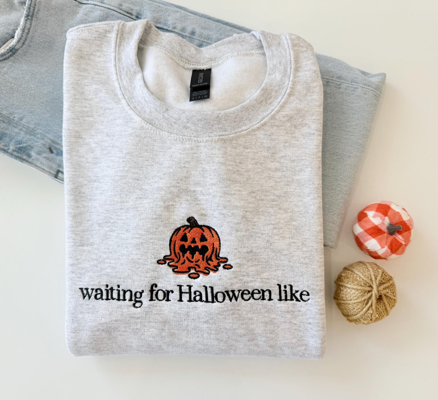 A light gray sweatshirt with an embroidered melting jack-o'-lantern and the text "waiting for Halloween like" is displayed on a light surface next to light wash jeans and two small decorative pumpkins.