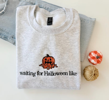 A light gray sweatshirt with an embroidered melting jack-o'-lantern and the text "waiting for Halloween like" is displayed on a light surface next to light wash jeans and two small decorative pumpkins.