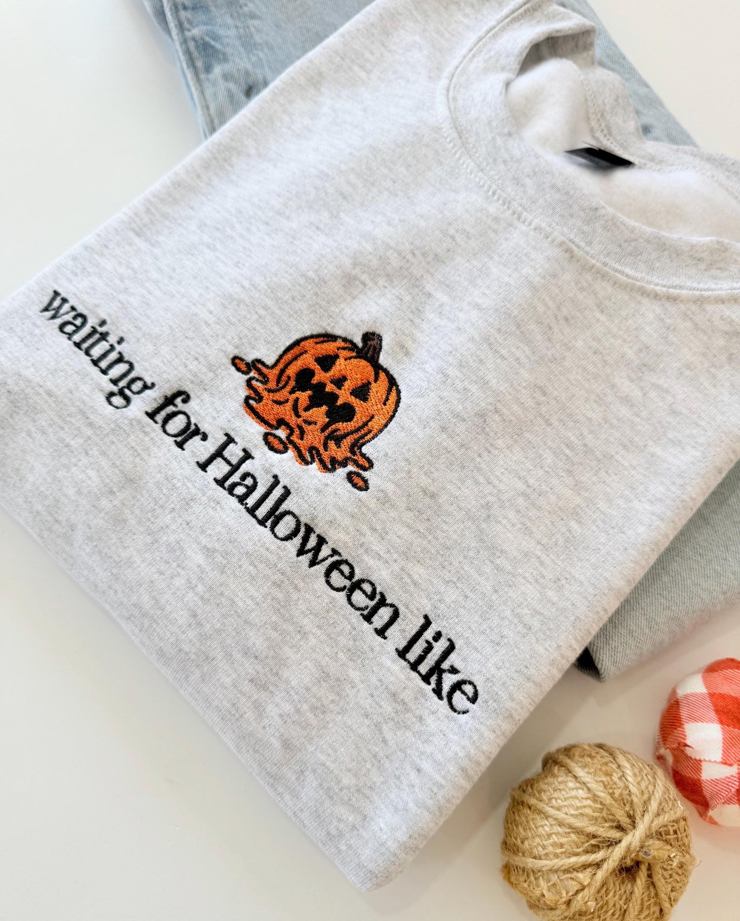 A light gray sweatshirt with an embroidered melting jack-o'-lantern and the text "waiting for Halloween like" is displayed on a light surface next to light wash jeans and two small decorative pumpkins.