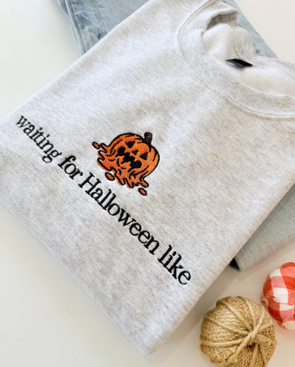 A light gray sweatshirt with an embroidered melting jack-o'-lantern and the text "waiting for Halloween like" is displayed on a light surface next to light wash jeans and two small decorative pumpkins.