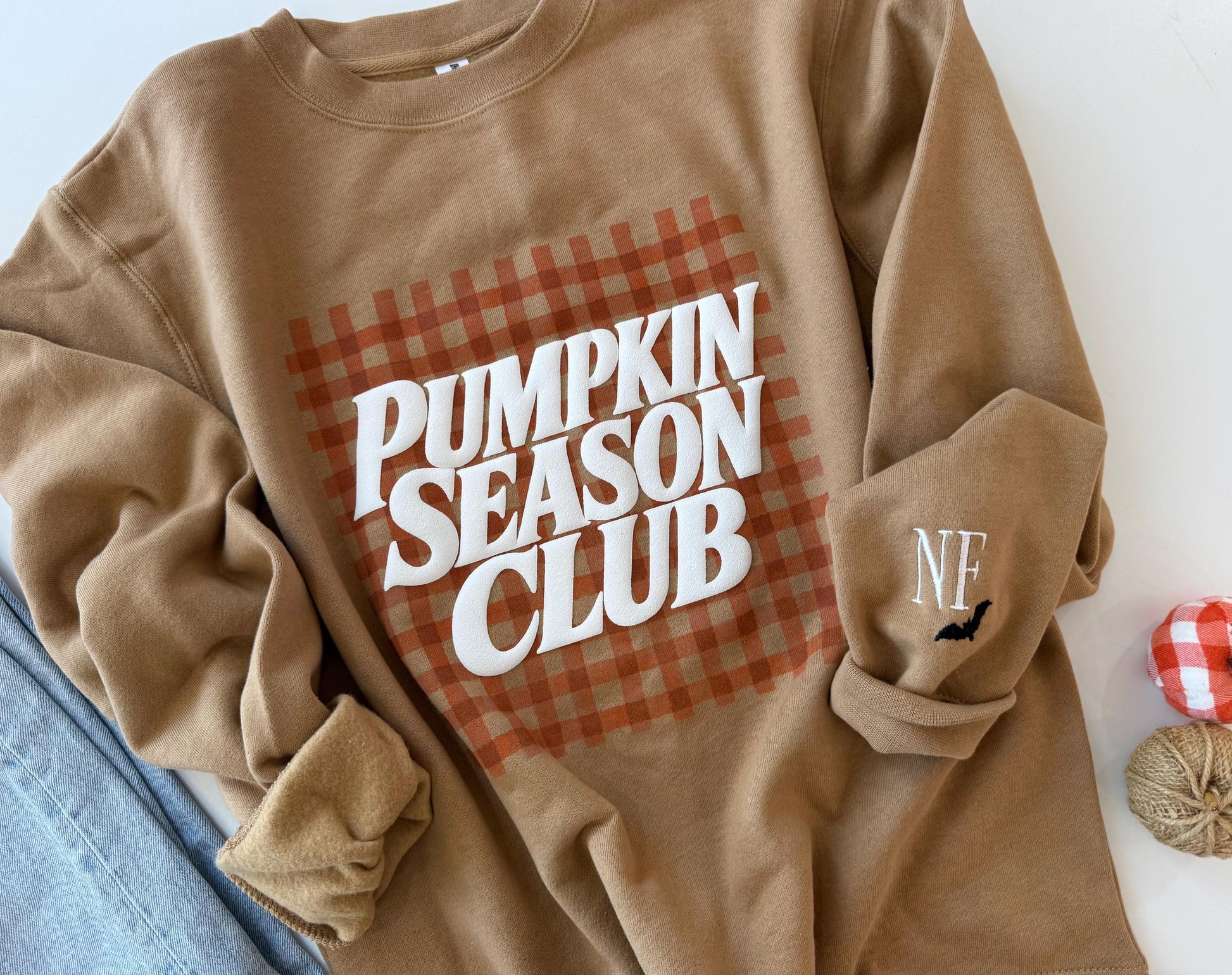 A tan sweatshirt with "PUMPKIN SEASON CLUB" in white text over an orange gingham pattern, and "NF" with a black bat embroidered on the left sleeve, next to blue jeans and fall decor.