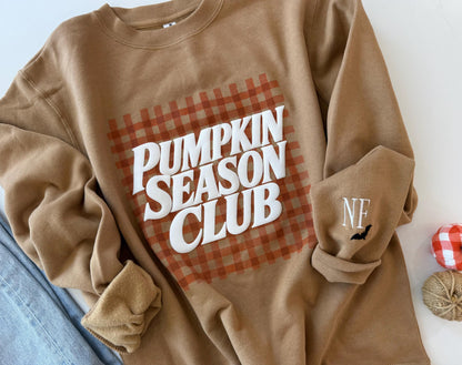 A tan sweatshirt with "PUMPKIN SEASON CLUB" in white text over an orange gingham pattern, and "NF" with a black bat embroidered on the left sleeve, next to blue jeans and fall decor.