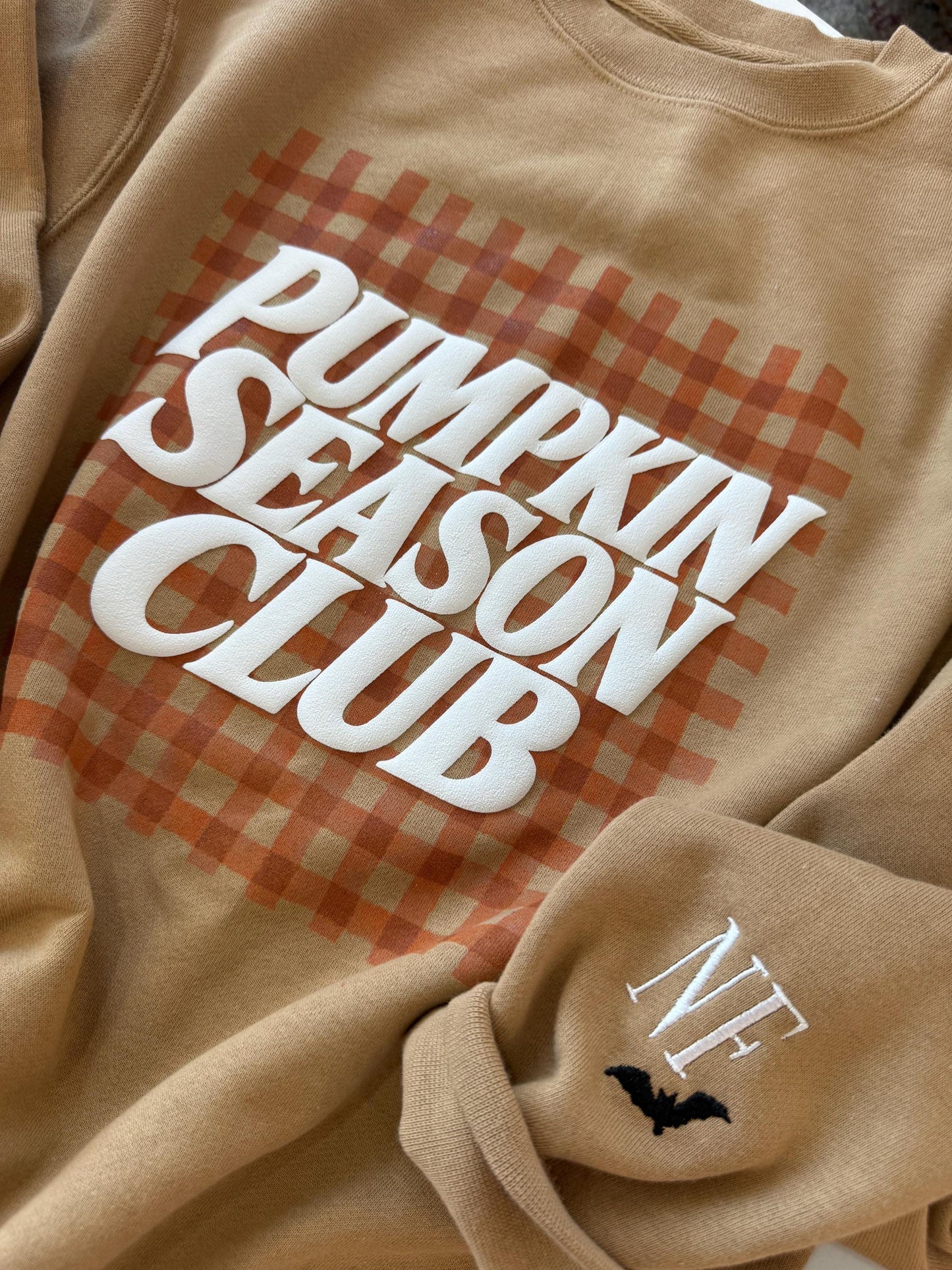 A tan sweatshirt with "PUMPKIN SEASON CLUB" in white text over an orange gingham pattern, and "NF" with a black bat embroidered on the left sleeve.