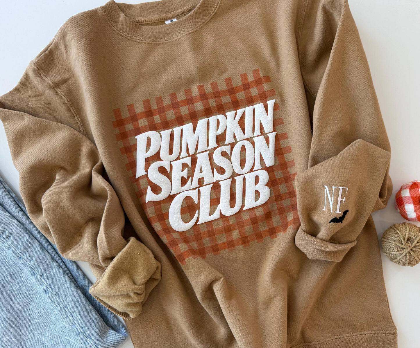 A tan sweatshirt with "PUMPKIN SEASON CLUB" in white text over an orange gingham pattern, and "NF" with a black bat embroidered on the left sleeve, next to blue jeans and fall decor.