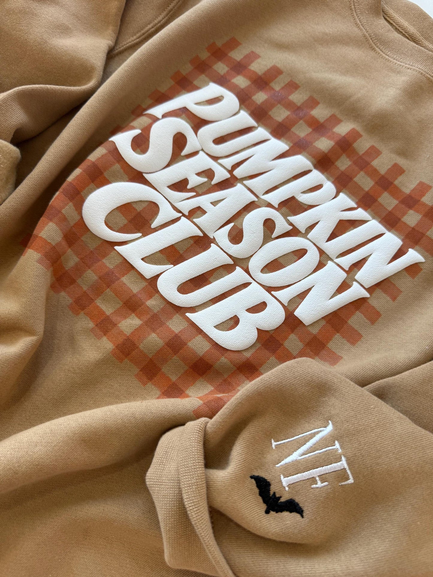 A close-up of a tan sweatshirt with "PUMPKIN SEASON CLUB" in white text over an orange gingham pattern, and "NF" with a black bat embroidered on the left sleeve.
