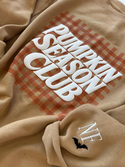 A close-up of a tan sweatshirt with "PUMPKIN SEASON CLUB" in white text over an orange gingham pattern, and "NF" with a black bat embroidered on the left sleeve.