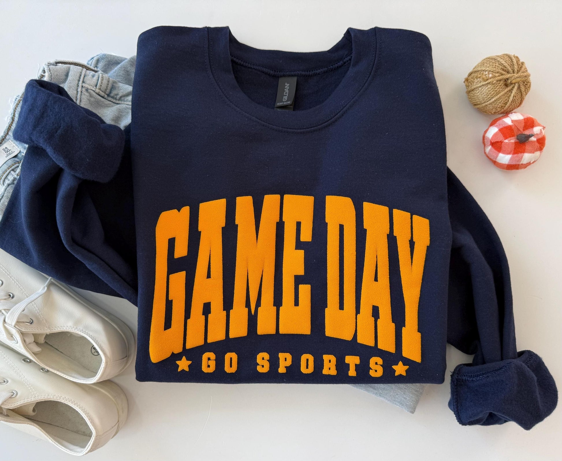 A navy blue crewneck sweatshirt with "GAME DAY" and "GO SPORTS" in orange embossed lettering, folded on a white surface next to light-wash jeans, white sneakers, and two small decorative gourds.