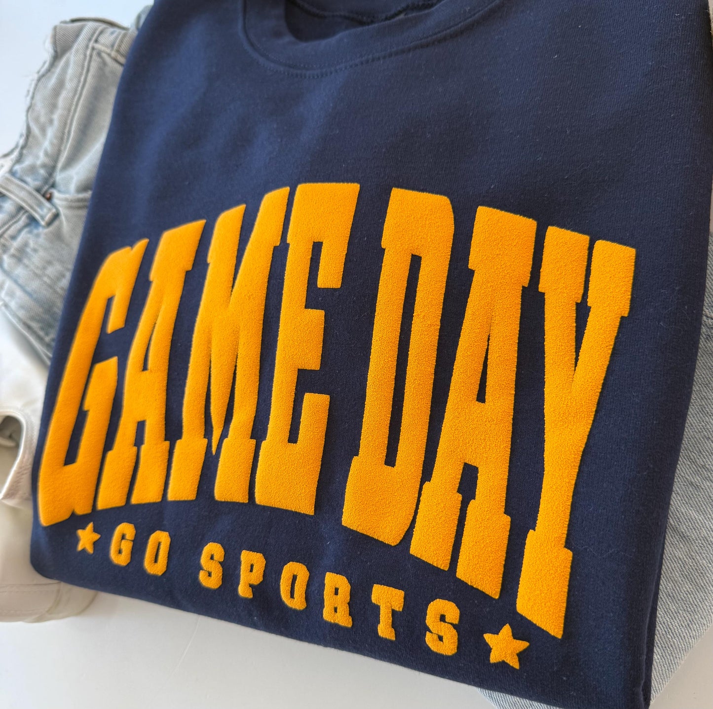 A close-up of a navy blue crewneck sweatshirt with "GAME DAY" and "GO SPORTS" in orange embossed lettering, folded on a white surface next to light-wash jeans.