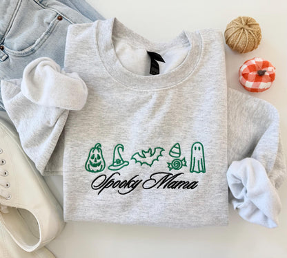 A light gray crewneck sweatshirt with green embroidered Halloween icons and "Spooky Mama" in black script, folded next to jeans and white sneakers.