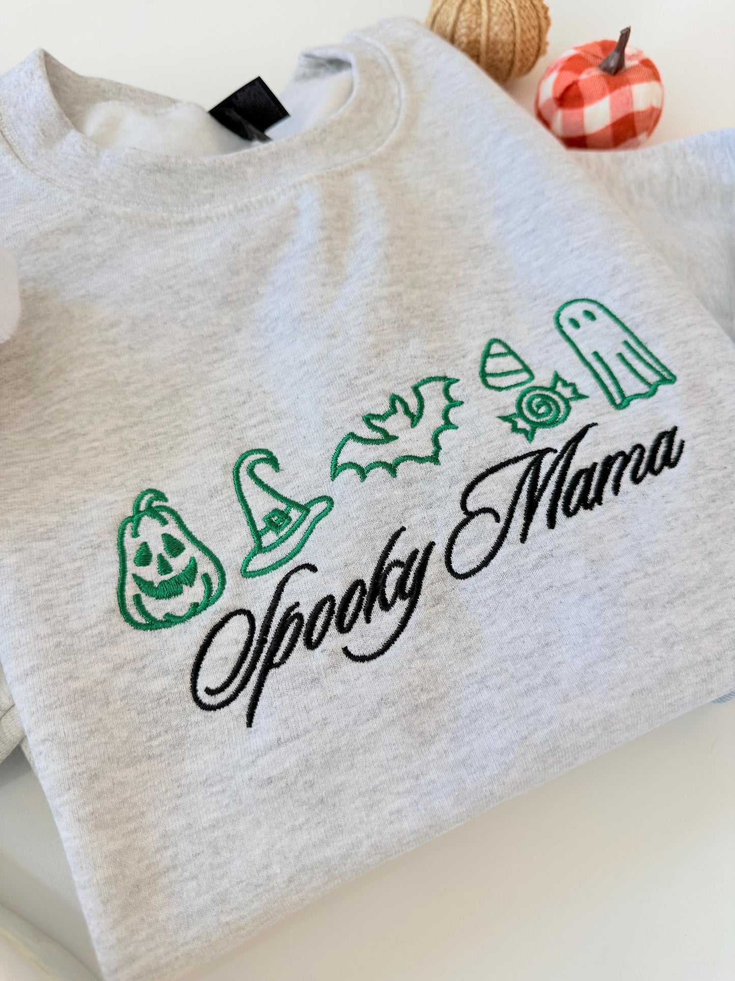 A close-up of a light gray crewneck sweatshirt with green embroidered Halloween icons and "Spooky Mama" in black script, with decorative pumpkins in the background.