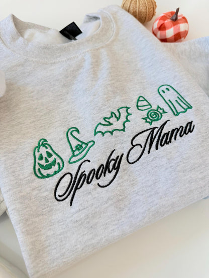 A close-up of a light gray crewneck sweatshirt with green embroidered Halloween icons and "Spooky Mama" in black script, with decorative pumpkins in the background.