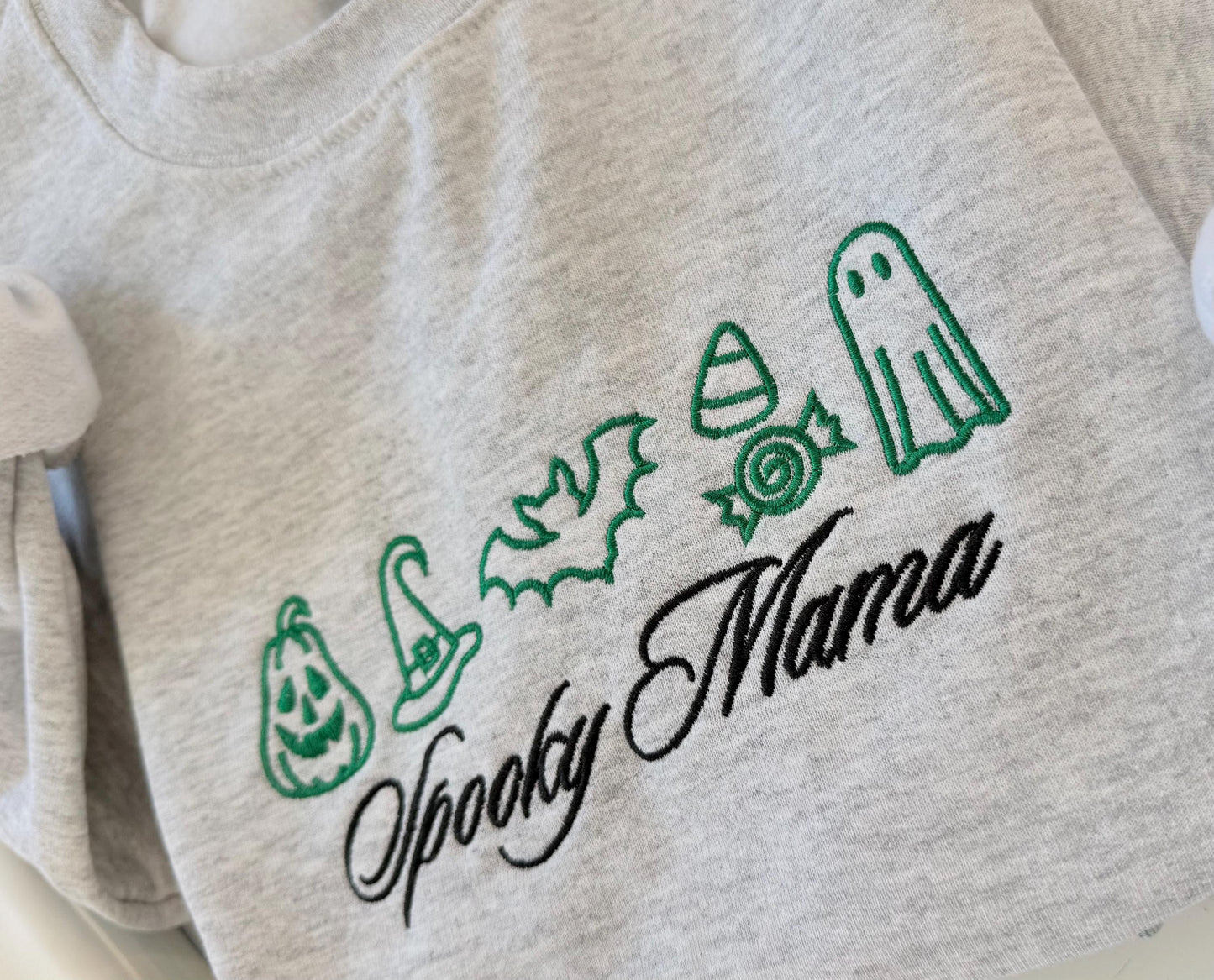 A close-up of the green embroidered Halloween icons including a pumpkin, witch hat, bat, candy corn, wrapped candy, and ghost, with "Spooky Mama" in black script on a gray sweatshirt.