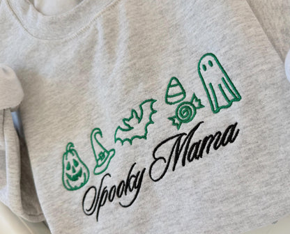 A close-up of the green embroidered Halloween icons including a pumpkin, witch hat, bat, candy corn, wrapped candy, and ghost, with "Spooky Mama" in black script on a gray sweatshirt.