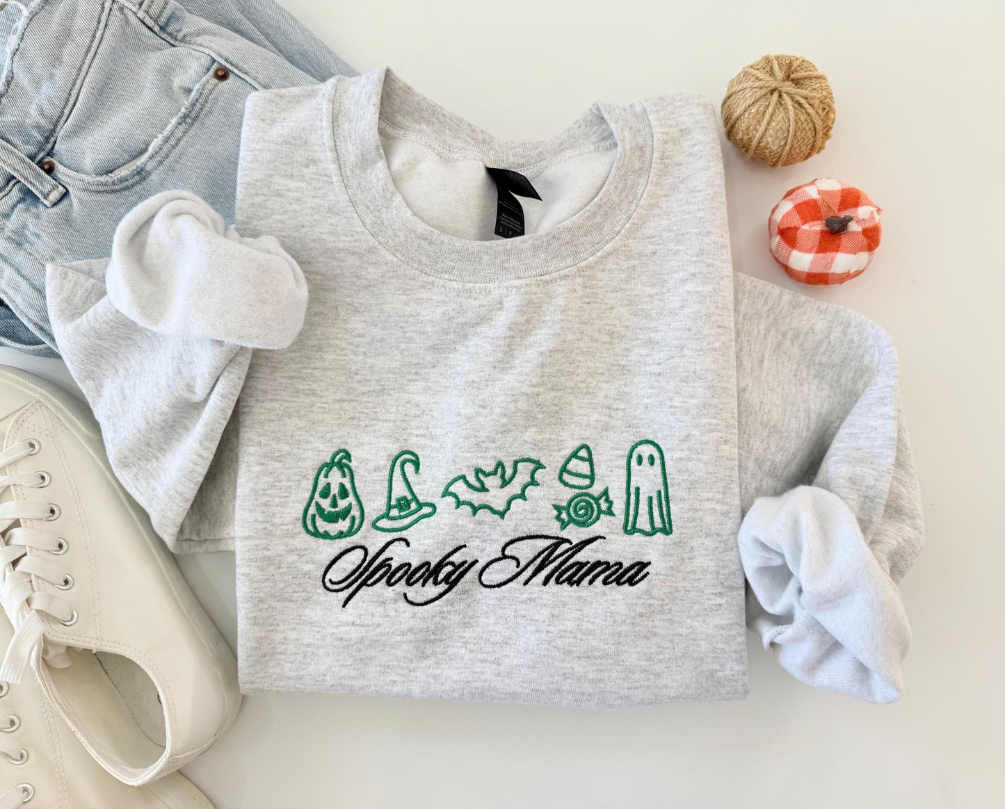 A light gray crewneck sweatshirt with green embroidered Halloween icons and "Spooky Mama" in black script, folded with jeans, white sneakers, and small decorative pumpkins.