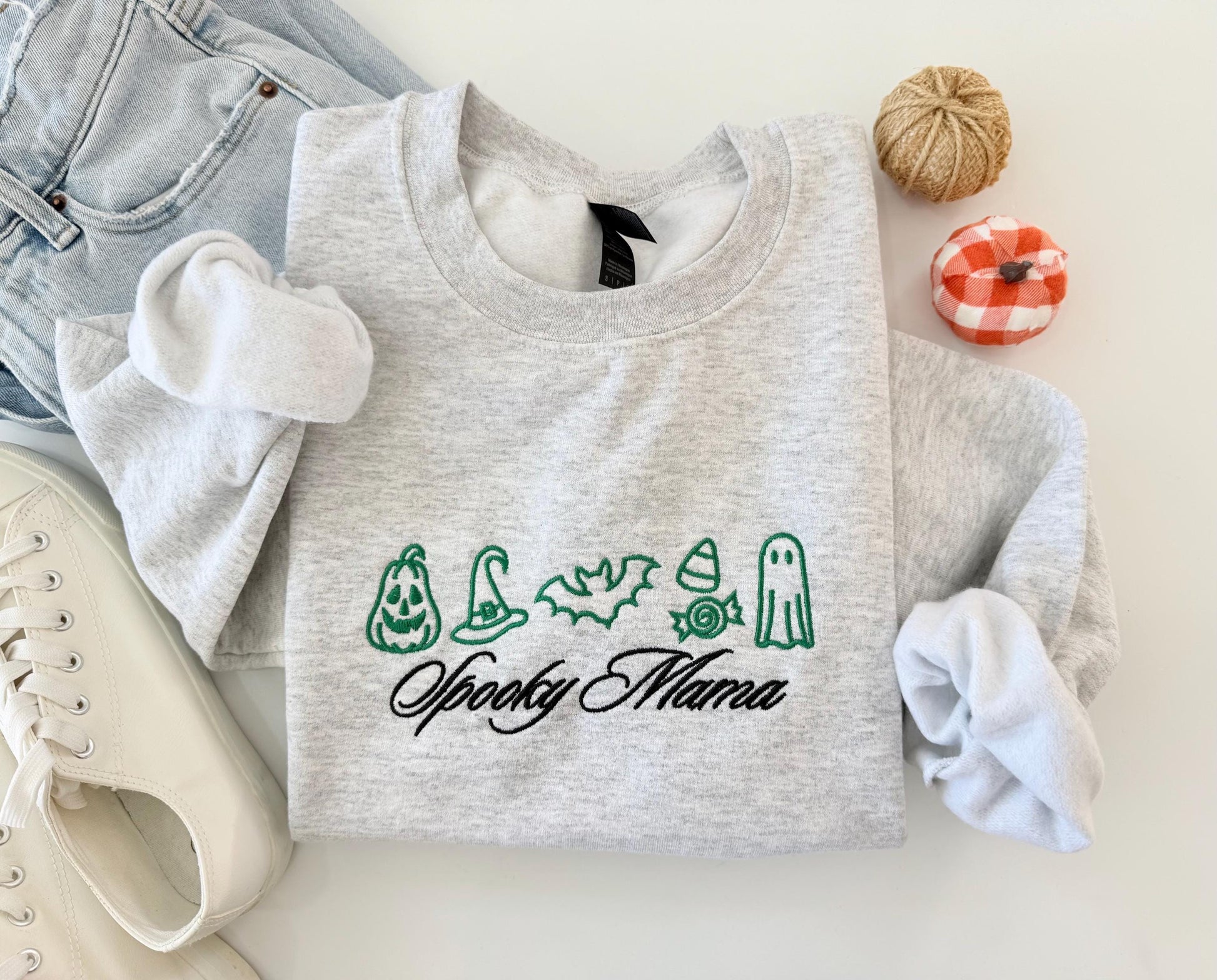 A light gray crewneck sweatshirt with green embroidered Halloween icons and "Spooky Mama" in black script, folded with jeans, white sneakers, and small decorative pumpkins.