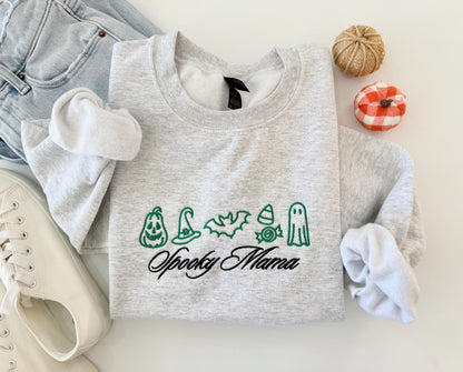 A light gray crewneck sweatshirt with green embroidered Halloween icons and "Spooky Mama" in black script, folded with jeans, white sneakers, and small decorative pumpkins.