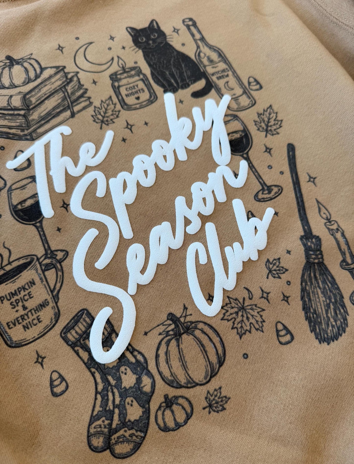 A close-up of a tan sweatshirt with "The Spooky Season Club" in white script, surrounded by black line drawings of Halloween and fall items, including a black cat, pumpkins, and a broom.