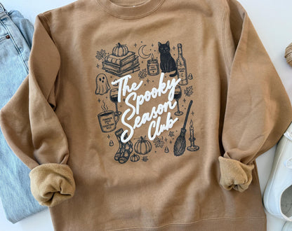 A tan crewneck sweatshirt with "The Spooky Season Club" in white script, surrounded by black line drawings of Halloween and fall items, including a black cat, pumpkins, and a broom, laid flat with rolled-up sleeves next to blue jeans and a white
