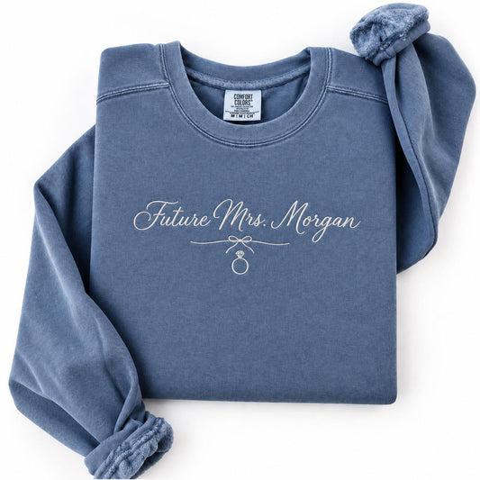Embroidered Future Mrs sweatshirt in Comfort Colors Blue Jean, flat lay showing personalized last name script and diamond ring stitched on left chest — engagement gift for the bride to be